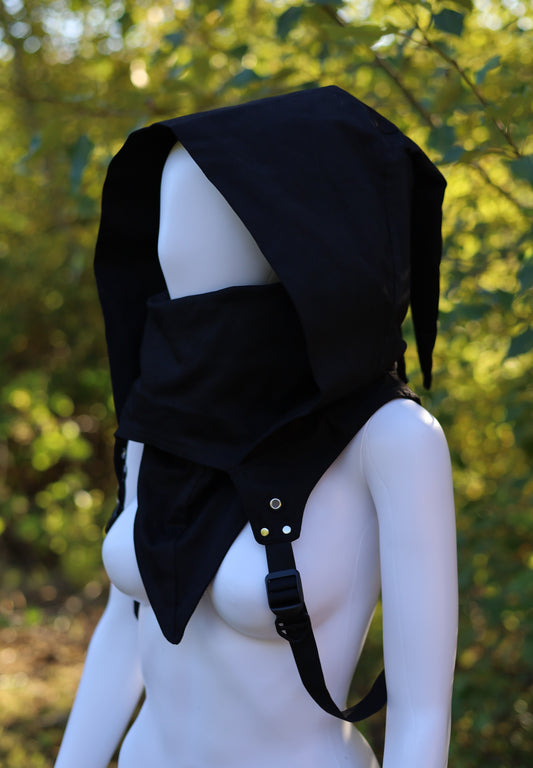 Mannequin wearing a black hooded garment with straps against a blurred green background