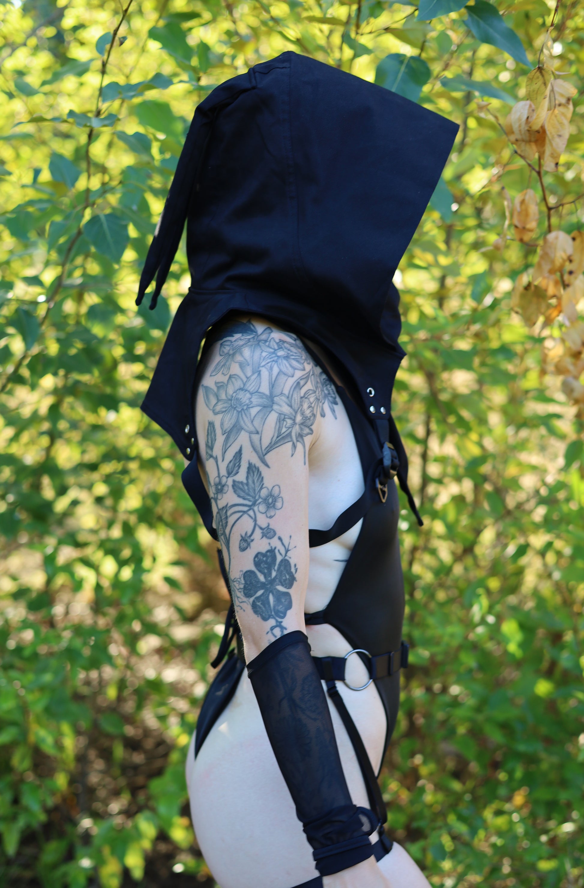 Person wearing a black hooded outfit with tattoos visible on arms against a natural background